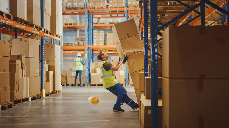 Shot of a Warehouse Worker Has Work Related Accident. He is Falling Down Before Trying to Pick Up Heavy Cardboard Box from the Shelf.